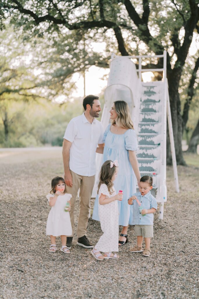 family photographers Colorado Springs captures parents hugging and smiling at each other while three kids play together in front of them