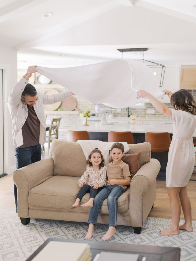 kids sit on couch while mom and dad spread a blanket over the couch like a tent