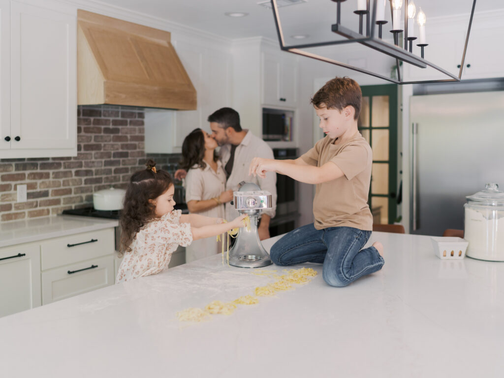 kids making pasta on kitchen island with parents kissing in background for Colorado in home family session