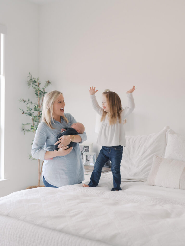 mom and newborn with sister jumping on bed