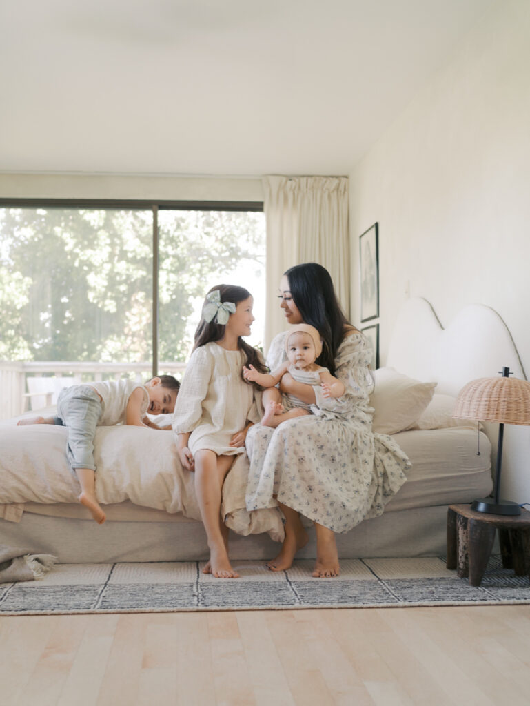 mom with oldest kid and youngest sitting on a bed interacting while another child climbs on bed