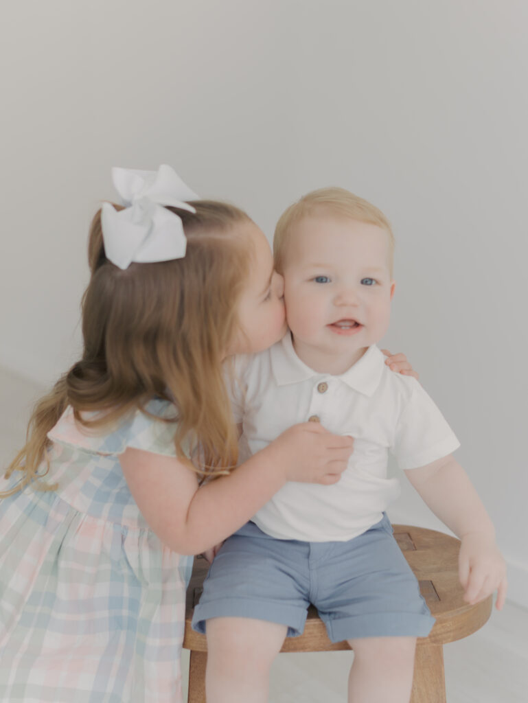 sister kissing little brothers cheek as he sits on stool for sibling mini sessions colorado springs