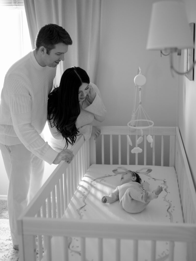 mom and dad leaning over crib looking at newborn baby for when to book newborn photos