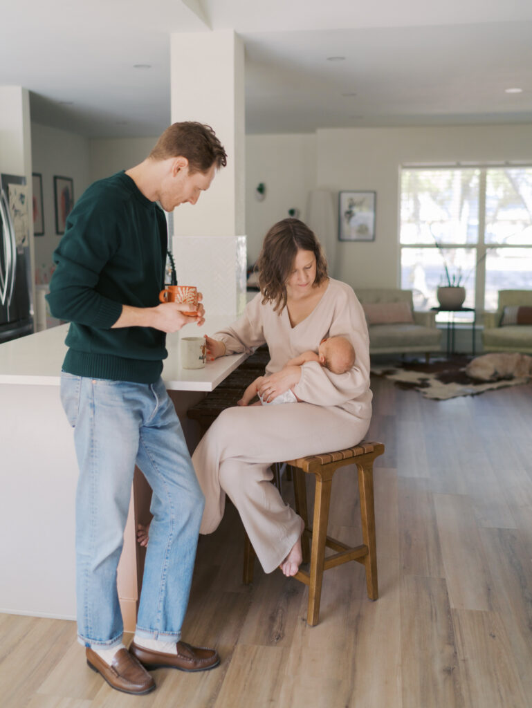 mom and dad enjoy coffee in kitchen while looking at newborn in mom's arms for Colorado Springs Newborn Photography